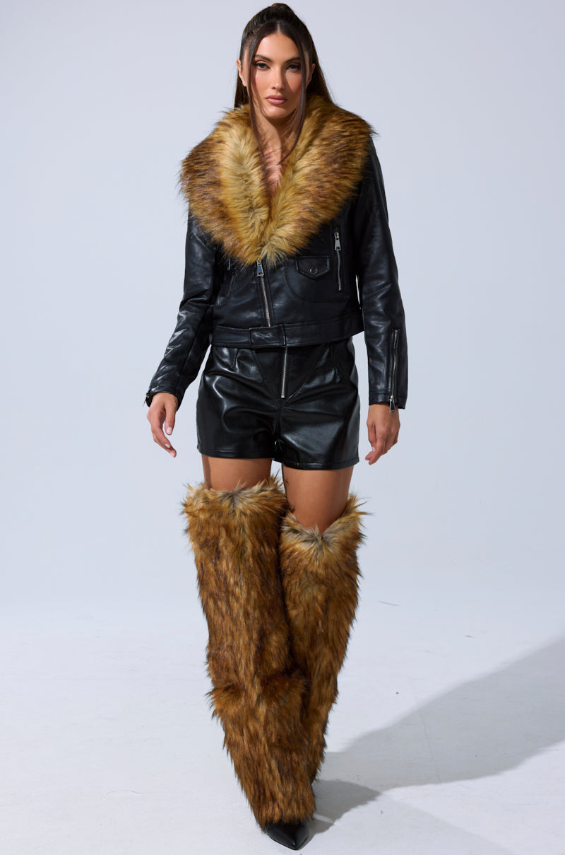 A woman models the AUSSY FUR COLLAR MOTO black faux leather jacket with a large fur collar, paired with black leather shorts and tall brown faux fur leg warmers—a striking statement in women’s outerwear.