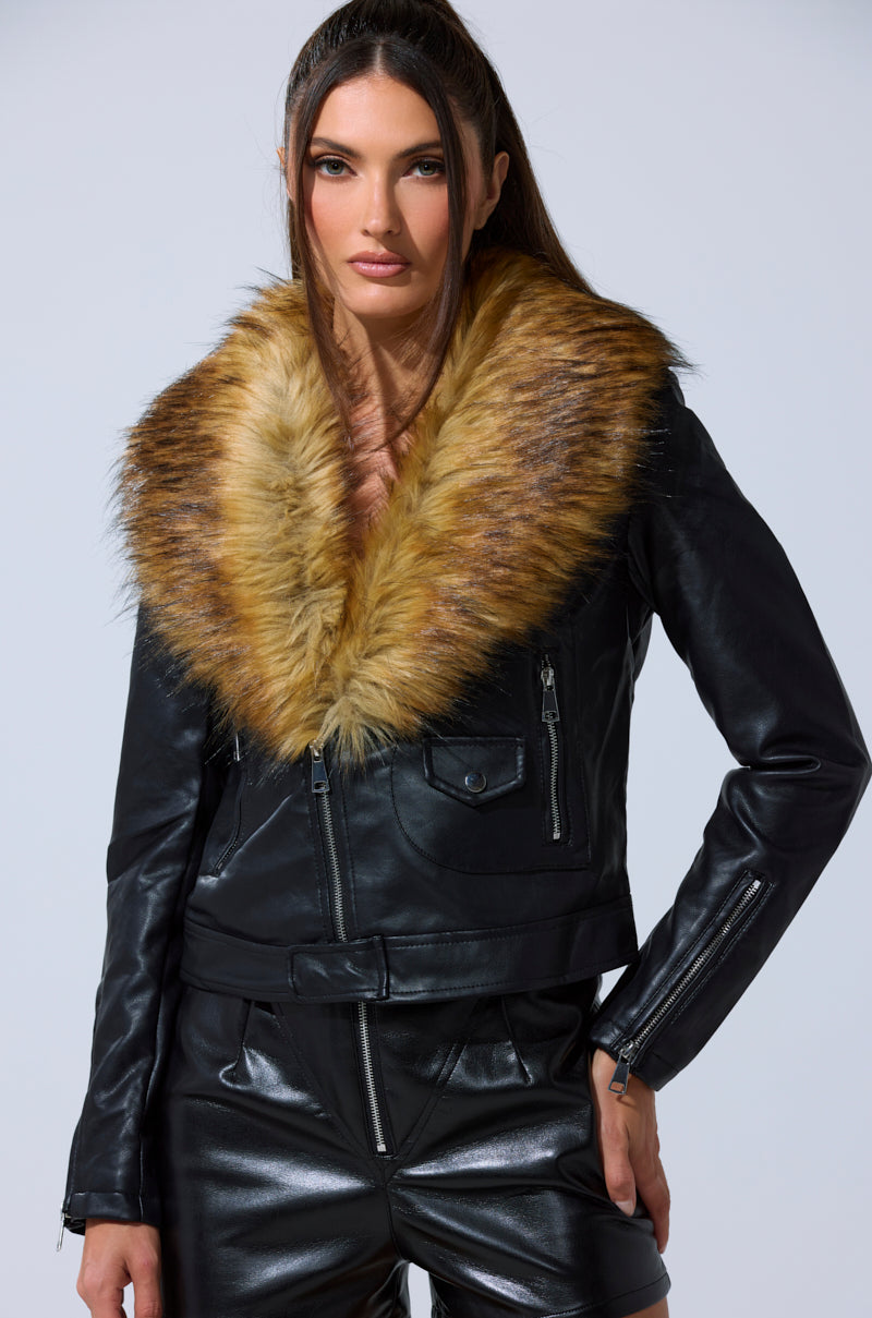 A woman with straight brown hair models the AUSSY FUR COLLAR MOTO, a black faux leather jacket with a large detachable fur collar, paired with black leather shorts against a plain light background.