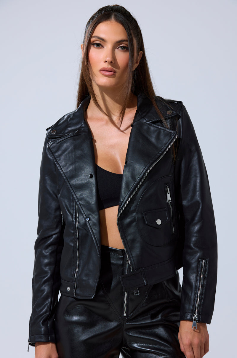 A woman wearing the AUSSY FUR COLLAR MOTO jacket over a black crop top and black pants stands against a plain light background, looking directly at the camera with a neutral expression.