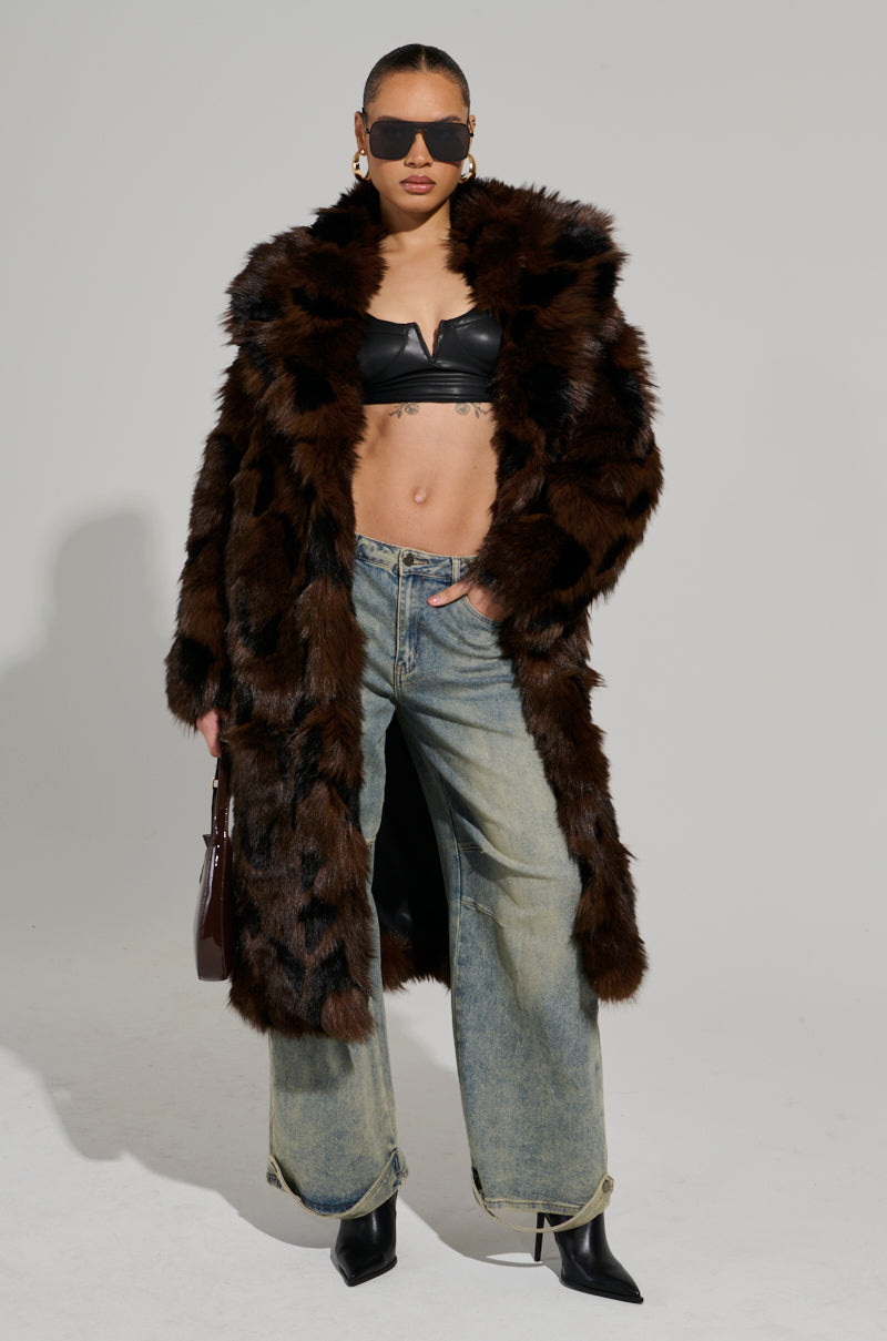 A person confidently stands wearing the AUSTIN FAUX FUR TRENCH at maxi-length over a black bralette, wide-legged jeans, and black pointed boots. They accessorize with dark sunglasses and carry a small handbag.