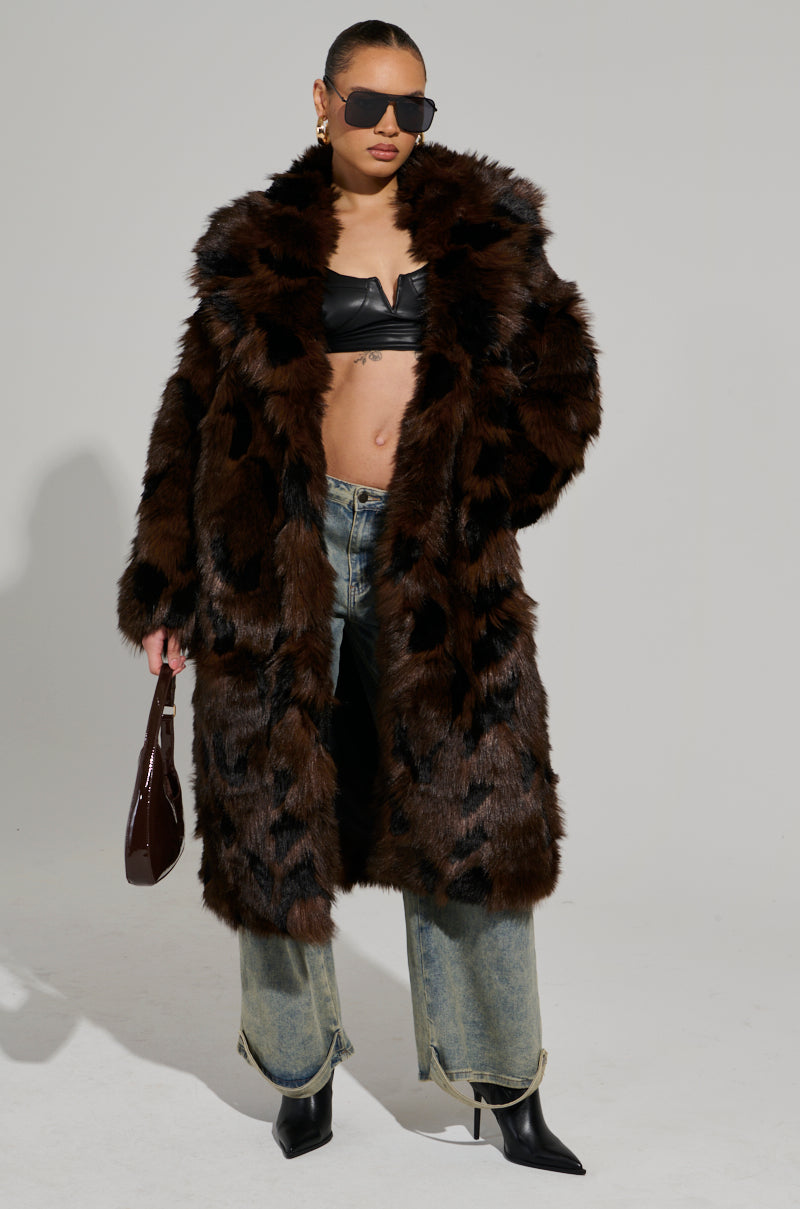 A person confidently poses in an AUSTIN FAUX FUR TRENCH, a maxi-length brown coat featuring a notch-lapel collar. They style it with sunglasses, a black top, jeans, a dark bag, and black heeled shoes against a plain background.