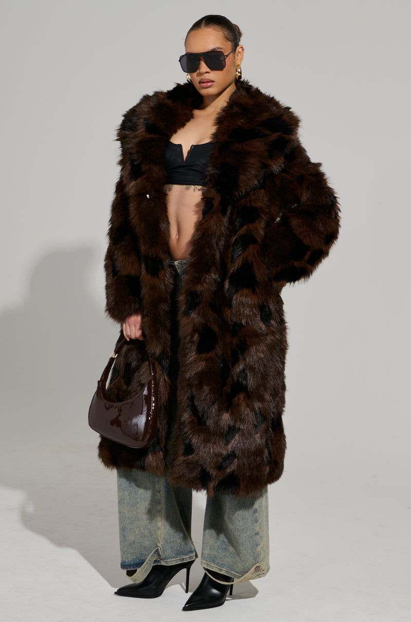 A person wearing oversized sunglasses, an AUSTIN FAUX FUR TRENCH with a notch-lapel collar, and blue jeans stands confidently. They hold a dark shiny handbag and wear black heeled boots against a plain background.