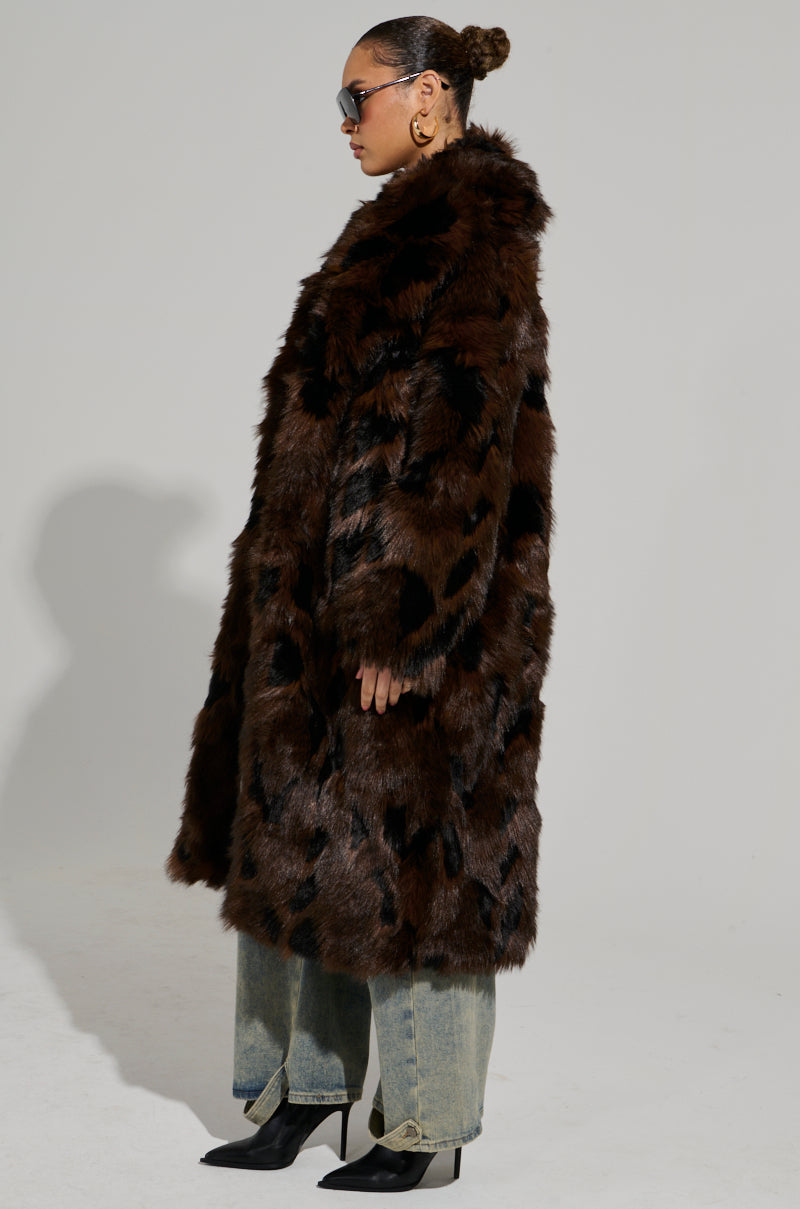 A person in the AUSTIN FAUX FUR TRENCH, featuring a maxi-length design with a chic black pattern on brown faux fur, stands in profile. Wearing sunglasses and gold hoop earrings, their hair is elegantly styled in a bun. The ensemble is completed with blue jeans and black heeled boots against the neutral backdrop.