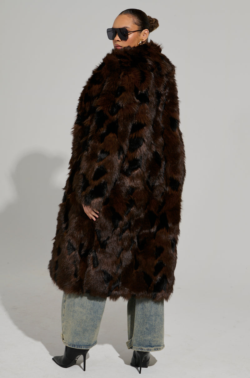 An individual confidently models the AUSTIN FAUX FUR TRENCH, featuring a maxi length and luxurious faux fur with distinct patterns. They accessorize with sunglasses, wide-leg jeans, and black high-heeled boots, radiating an effortlessly chic vibe.