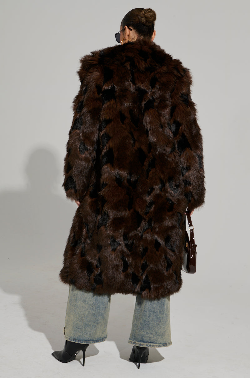 A person with hair in a bun stands with their back to the camera, wearing the AUSTIN FAUX FUR TRENCH—a long, dark brown coat featuring a notch-lapel collar. The ensemble is completed by wide-leg jeans, black pointed boots, sunglasses, and a small bag, while the patterned texture of the trench coat adds intrigue.