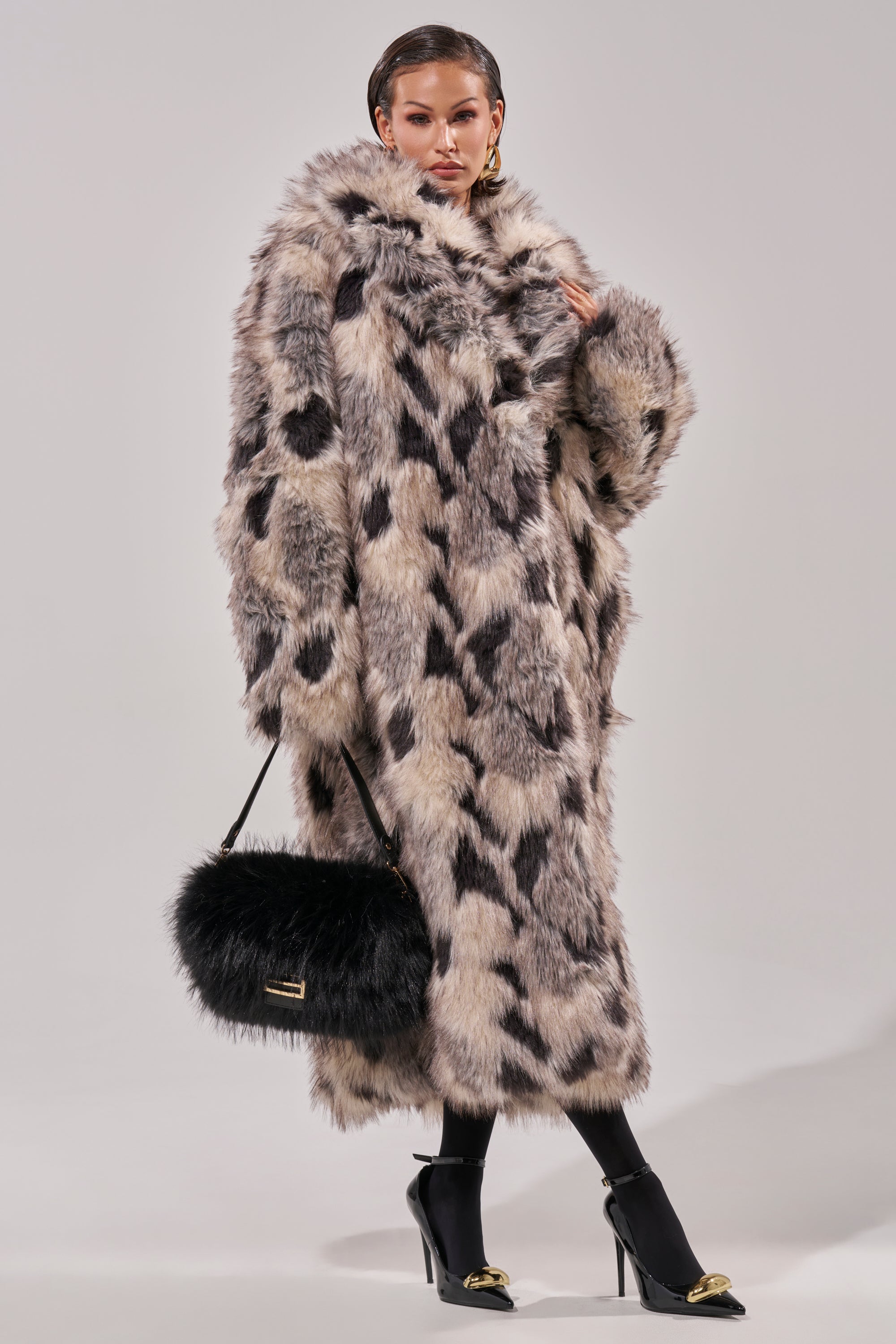 Wearing the NEW AUSTIN FAUX FUR COAT in an oversized, black and white spotted design, this person exudes vintage luxury paired with a black furry handbag, gold-accented black heels, slicked-back hair, and bold earrings.