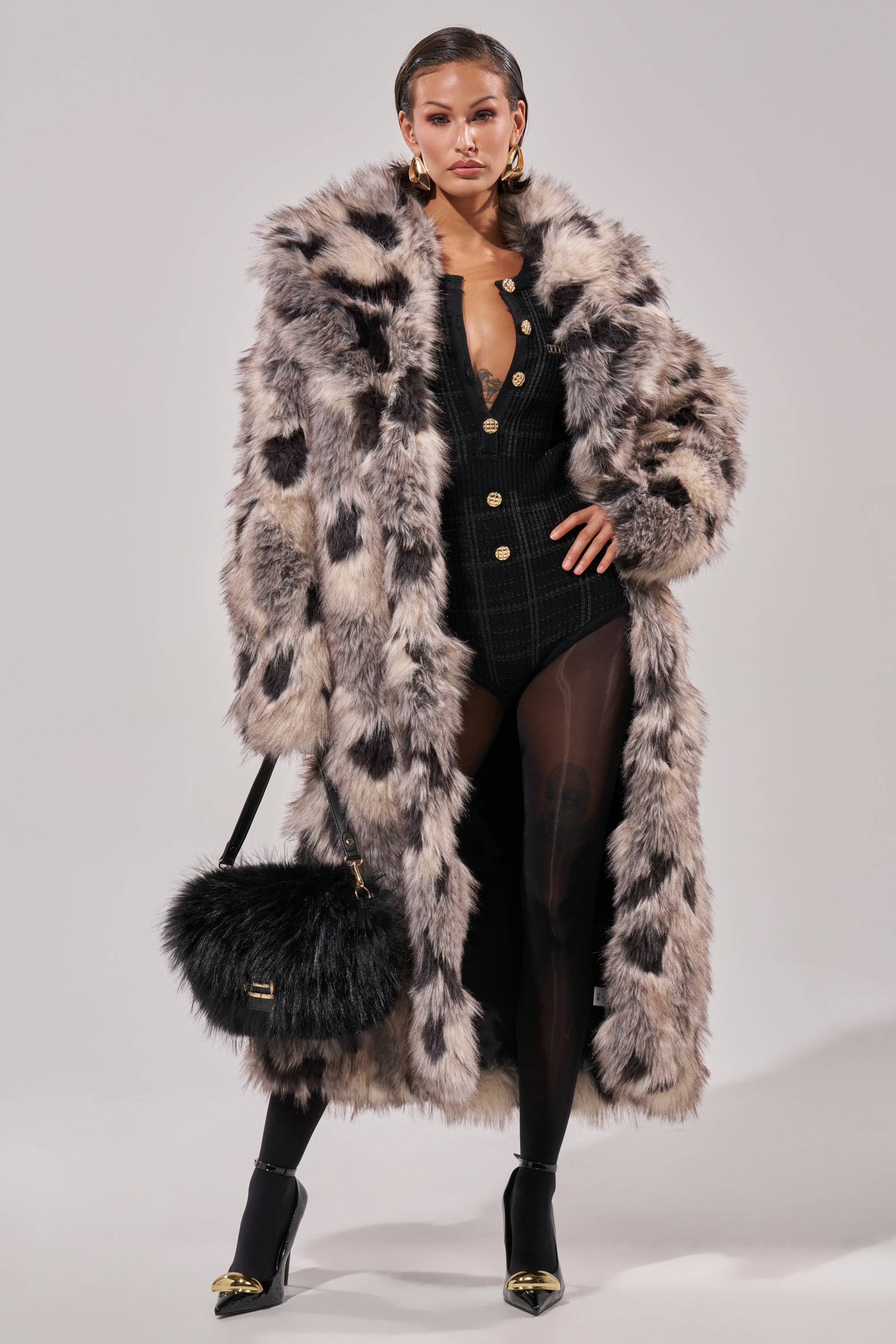 A woman stands confidently in the NEW AUSTIN FAUX FUR COAT with black and white spots over a black buttoned bodysuit, sheer tights, high heels, gold earrings, and holds a furry black handbag.