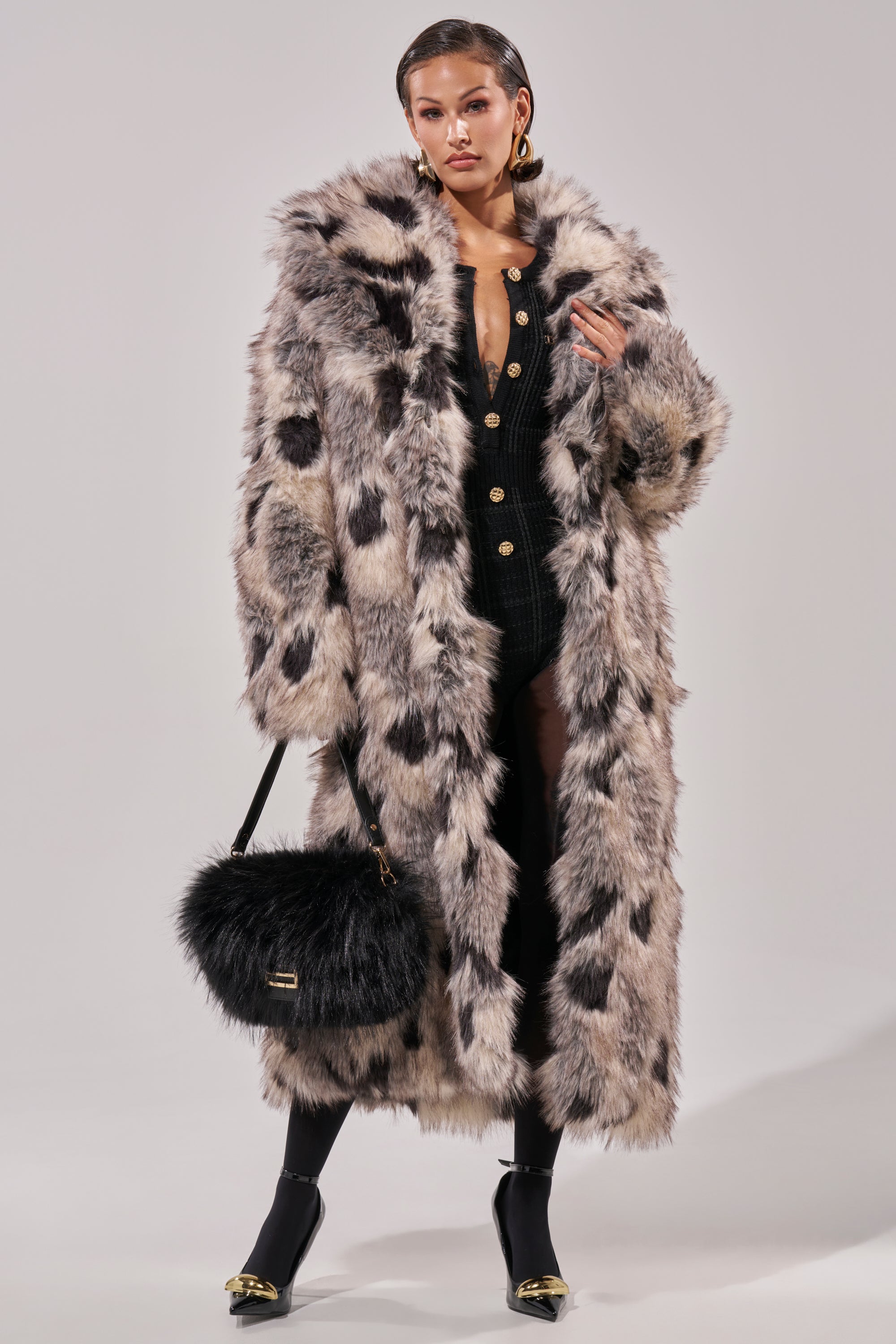Wearing the NEW AUSTIN FAUX FUR COAT with black and white spots, a black outfit with gold buttons, black heels, and a fluffy black handbag, a person stands against a plain light background.