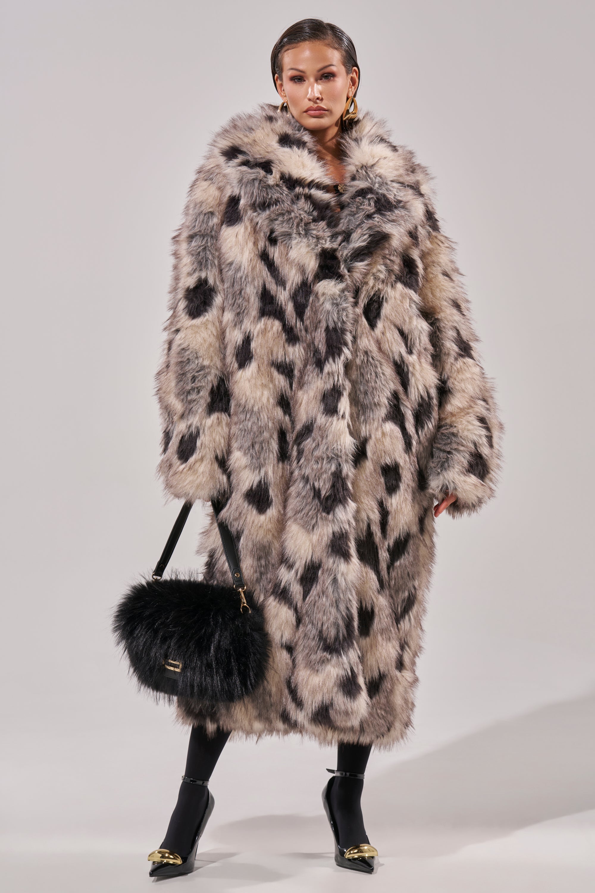 A woman stands confidently in the NEW AUSTIN FAUX FUR COAT with a black and white spotted pattern, styled with black tights, gold-detailed heels, and a black furry handbag against a plain light gray background.