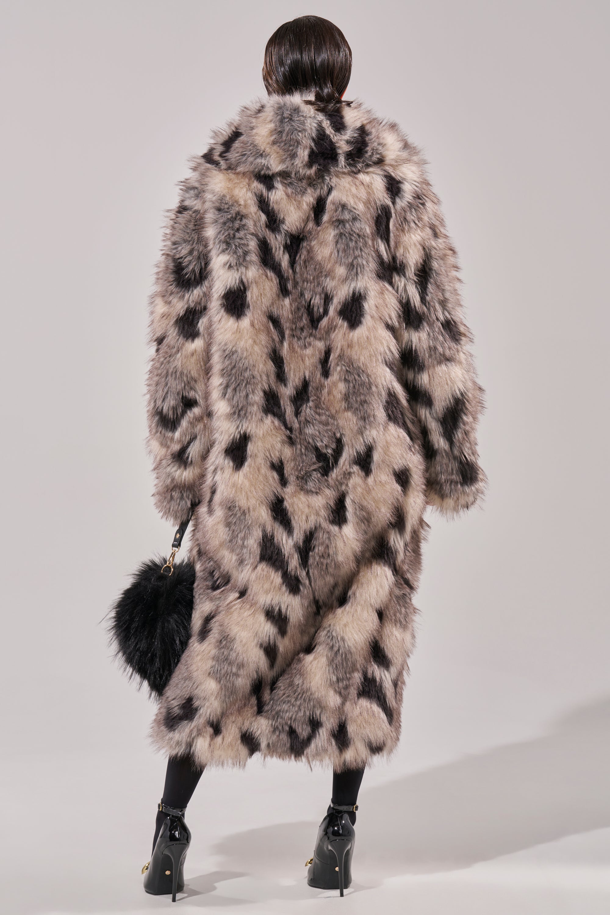 With their back to the camera, a person wears the NEW AUSTIN FAUX FUR COAT, accessorized with a matching black faux fur handbag, black heeled shoes, and tights.