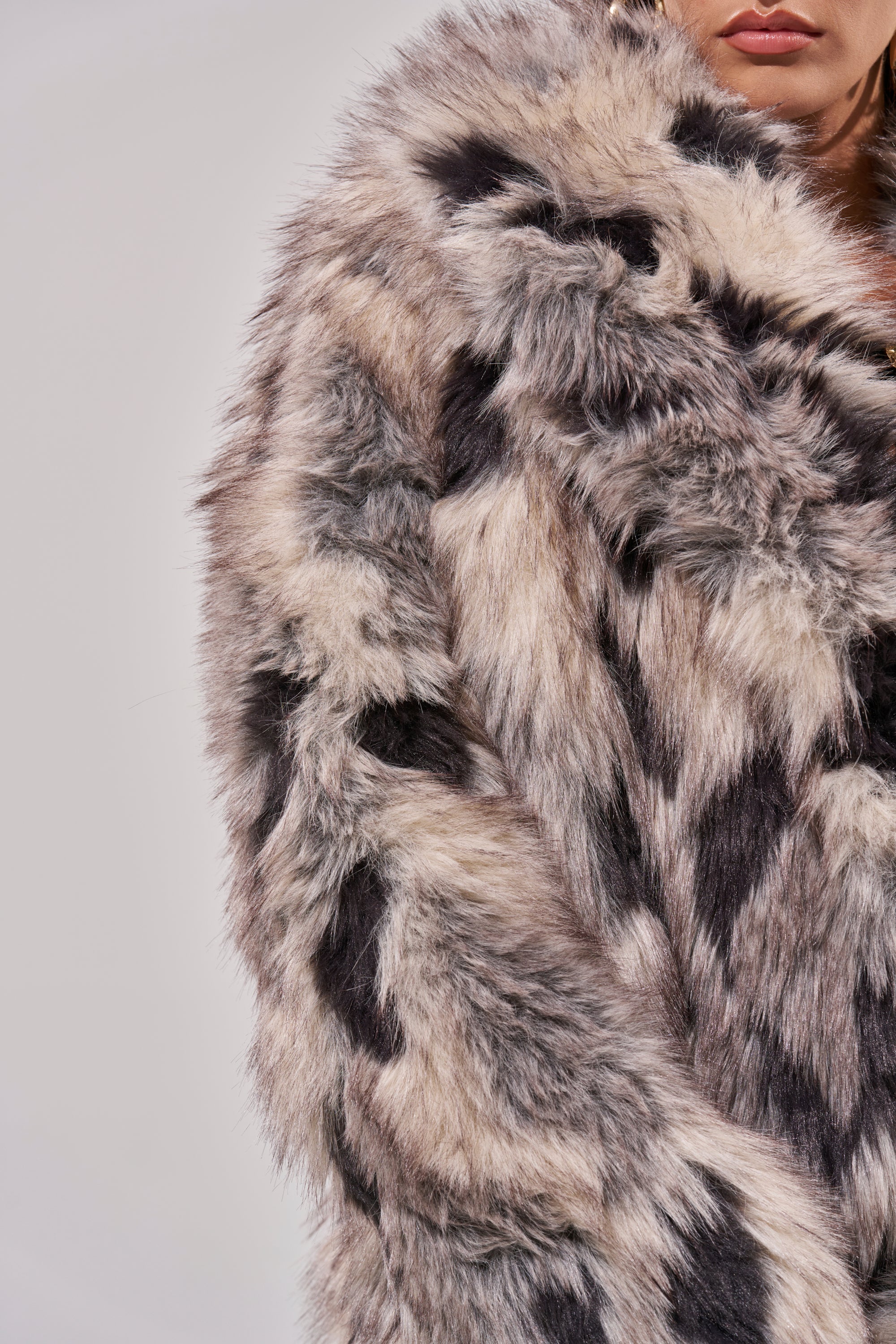 Wearing the NEW AUSTIN FAUX FUR COAT in gray, black, and beige tones, a person is photographed from the chest up against a plain background with only part of their face visible.