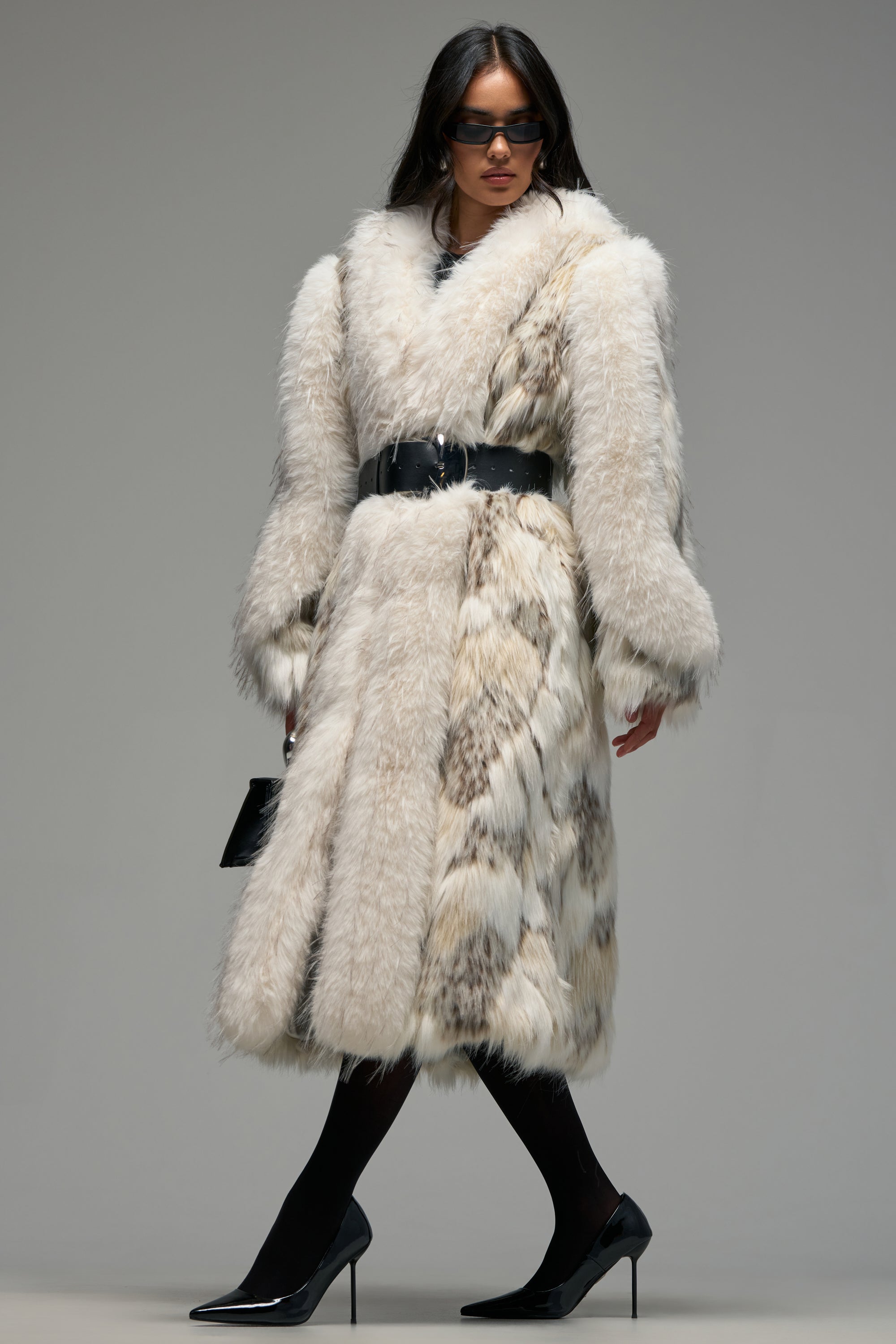 Wearing the GET TO IT PATCHWORK FAUX FUR coat—an elegant, long, ivory faux fur piece accented with a wide black belt—a woman pairs it with black high heels, black tights, bold sunglasses, and a small black purse to exemplify flawless winter style against a minimalist gray backdrop.