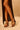 A person in a black high-slit dress showcases their legs and wears AZALEA WANG AVENA BLACK PAT SANDAL—black vegan leather heels with thin straps and a square toe—while standing against a plain beige background.