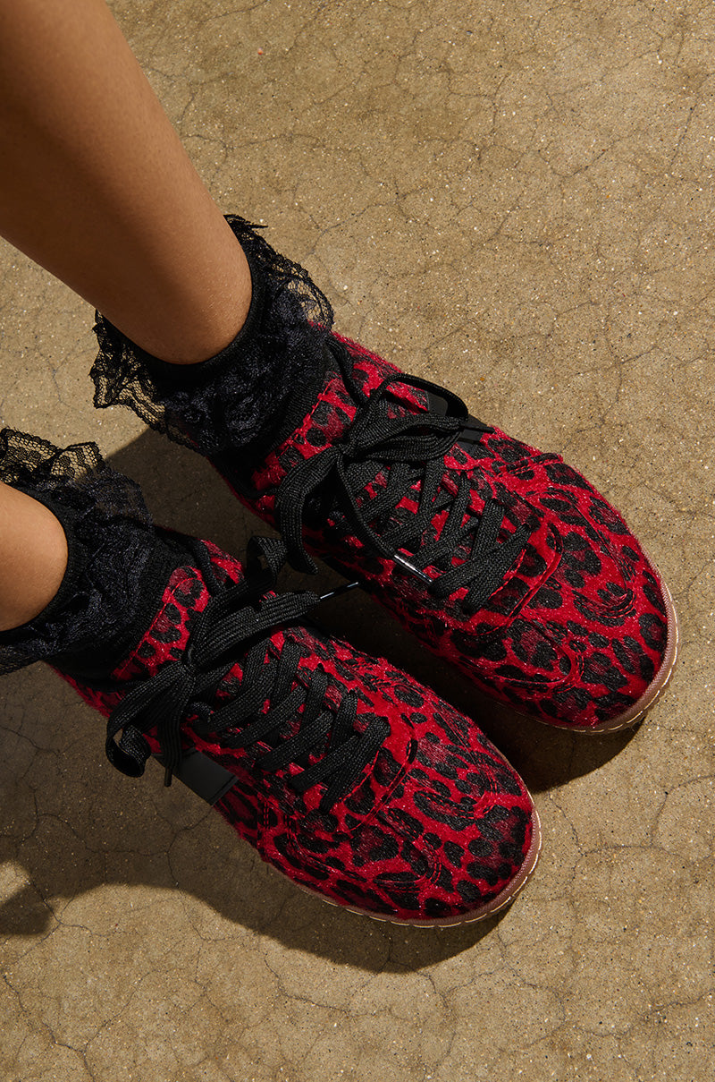 A person wearing the AZALEA WANG AVENGER RED LEOPARD SNEAKER with a black lace-up vamp and faux pony hair stands on a cracked concrete surface, complemented by their stylish black lace ankle socks.