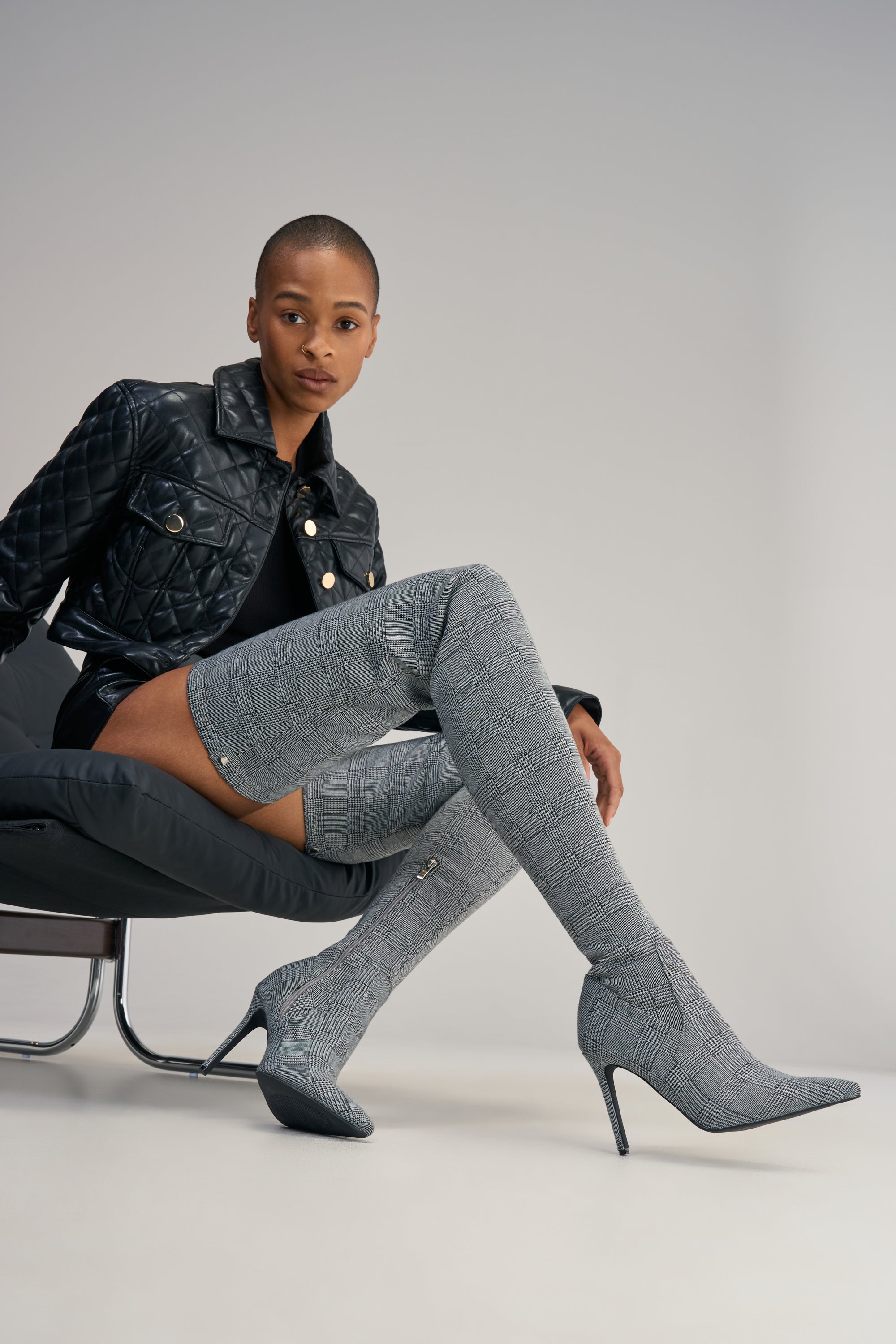 A person sits on a chair wearing a black quilted jacket, black shorts, and the AZALEA WANG AVENOIR HOUNDSTOOTH OVER THE KNEE FOUR WAY STRETCH BOOT. The background is plain and light-colored.