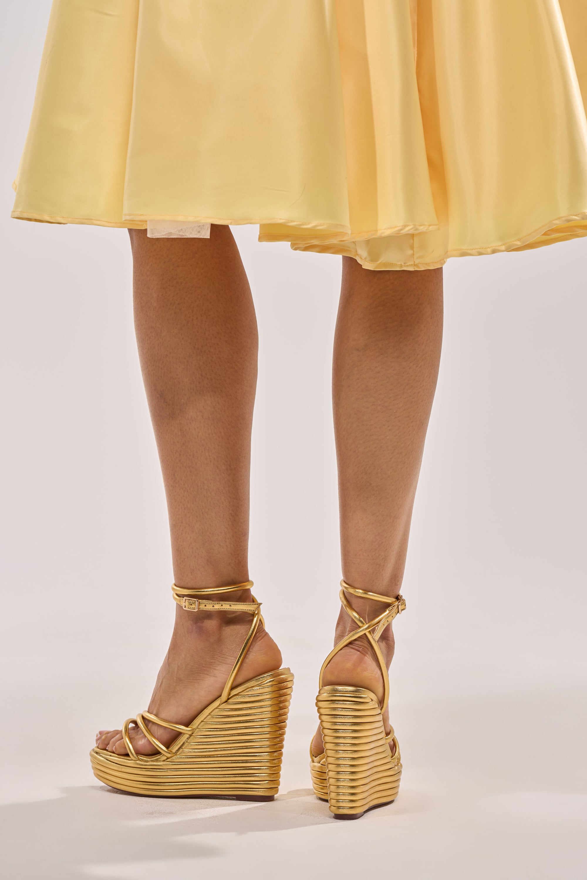 A person wearing a yellow, knee-length skirt and the AZALEA WANG AVENY GOLD STRAP ROUND TOE WEDGE SANDAL stands on a plain light background, shown from the knees down—an elegant choice for occasion wear. 2