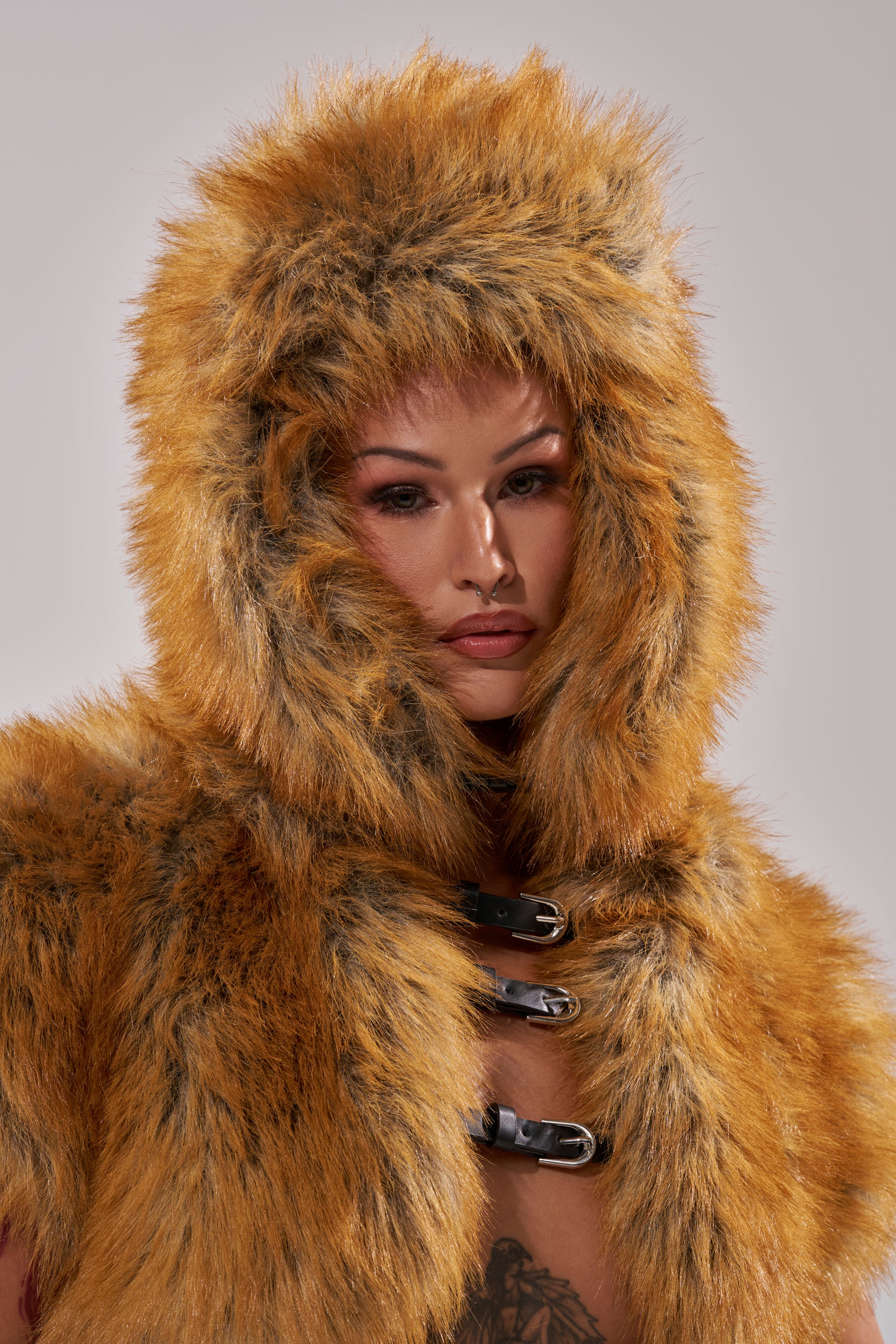 A person wears the SO WITH IT FAUX FUR AVIATOR HAT in orange and brown faux fur with dark streaks, featuring a hood framing their face, three black front buckles, and a buckle chin strap.