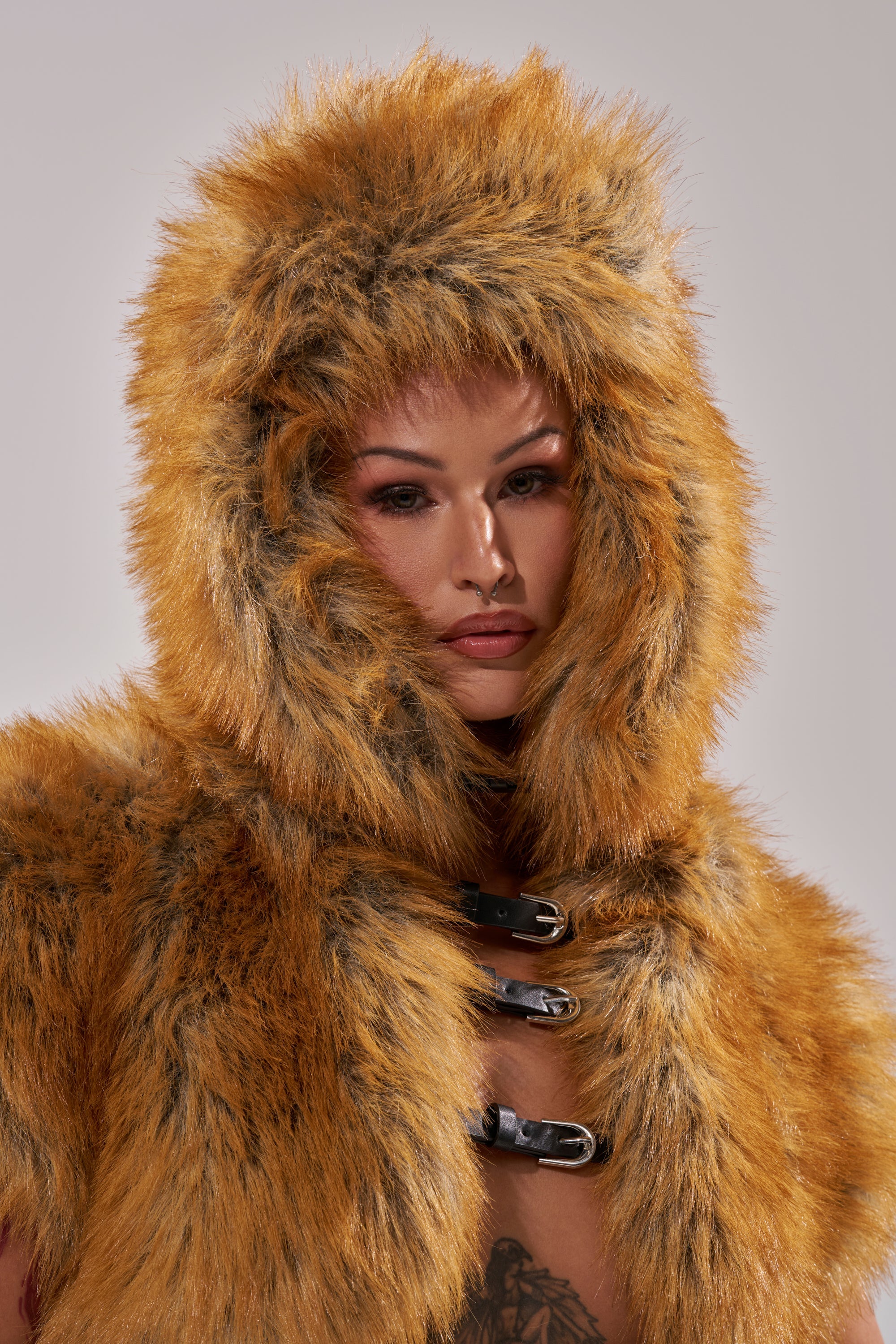 A person wears the SO WITH IT FAUX FUR AVIATOR HAT in orange and brown faux fur with dark streaks, featuring a hood framing their face, three black front buckles, and a buckle chin strap. 7