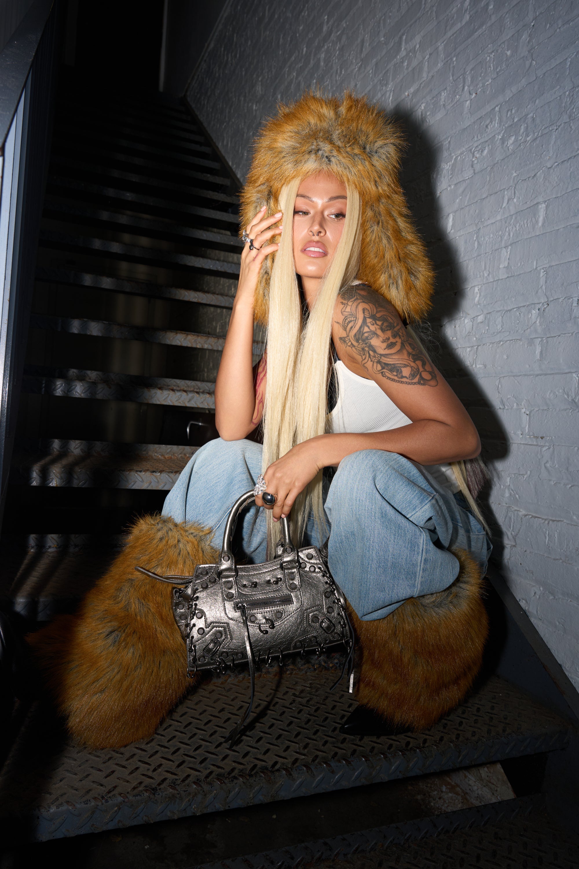 A woman with long blonde hair, tattoos, and jeans sits on metal stairs, wearing the SO WITH IT FAUX FUR AVIATOR HAT, matching boots, a white tank top, and holding a silver handbag as she poses thoughtfully. 3