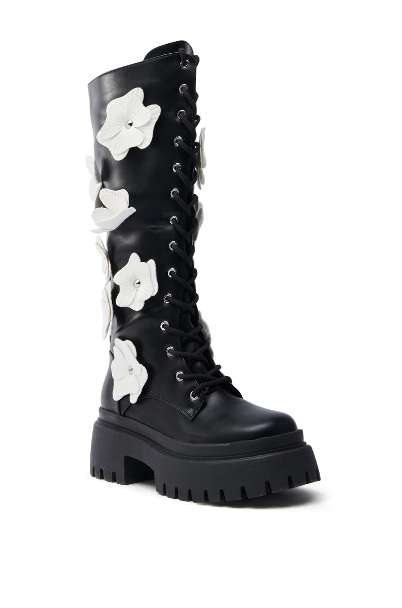The AZALEA WANG MIRABELLA BLACK FLORAL EMBELLISHED BOOT is a black knee-high combat boot with a chunky sole, featuring large white flower appliques on the side and faux leather lace-up details for a bold, stylish look.