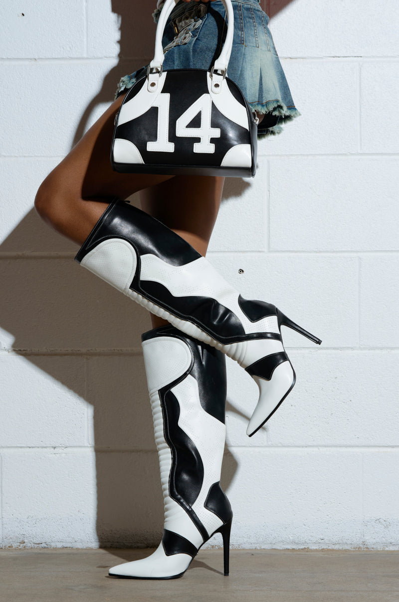 A person wearing AZALEA WANG RODRIGO Black and White Stiletto Boots leans against a white brick wall, holding a matching black and white handbag with the number "14" while donning a denim skirt.