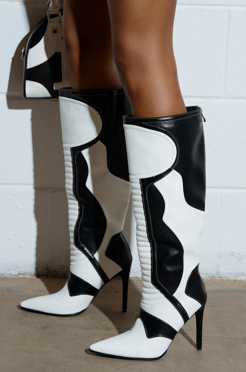 A person is wearing the AZALEA WANG RODRIGO Black and White Boot Stiletto, knee-high with a geometric design and pointed toes, made from vegan leather. These boots are styled with a matching handbag against a white textured wall and concrete floor backdrop.