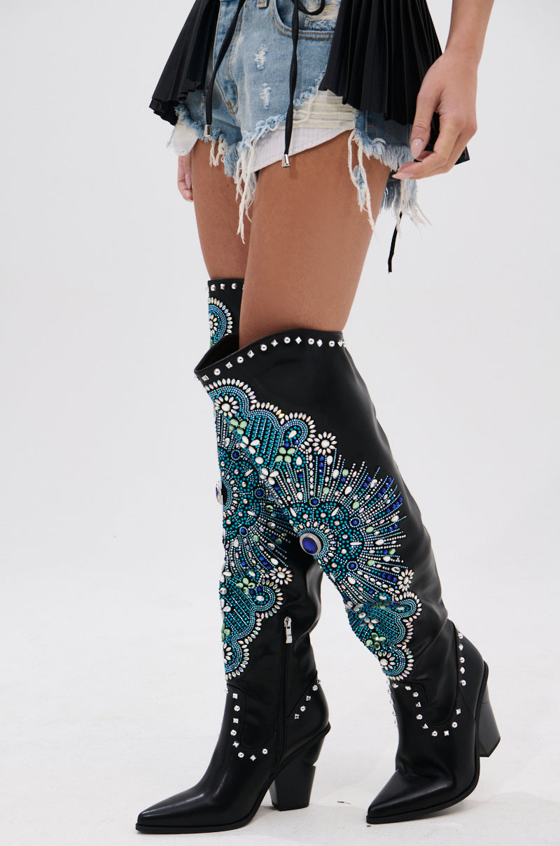 A person wears distressed denim shorts, a black pleated top, and the AZALEA WANG AXELBEAT BLACK EMBELLISHED WESTERN BOOT adorned with rhinestones, blue, green, and silver beadwork, plus stud accents.