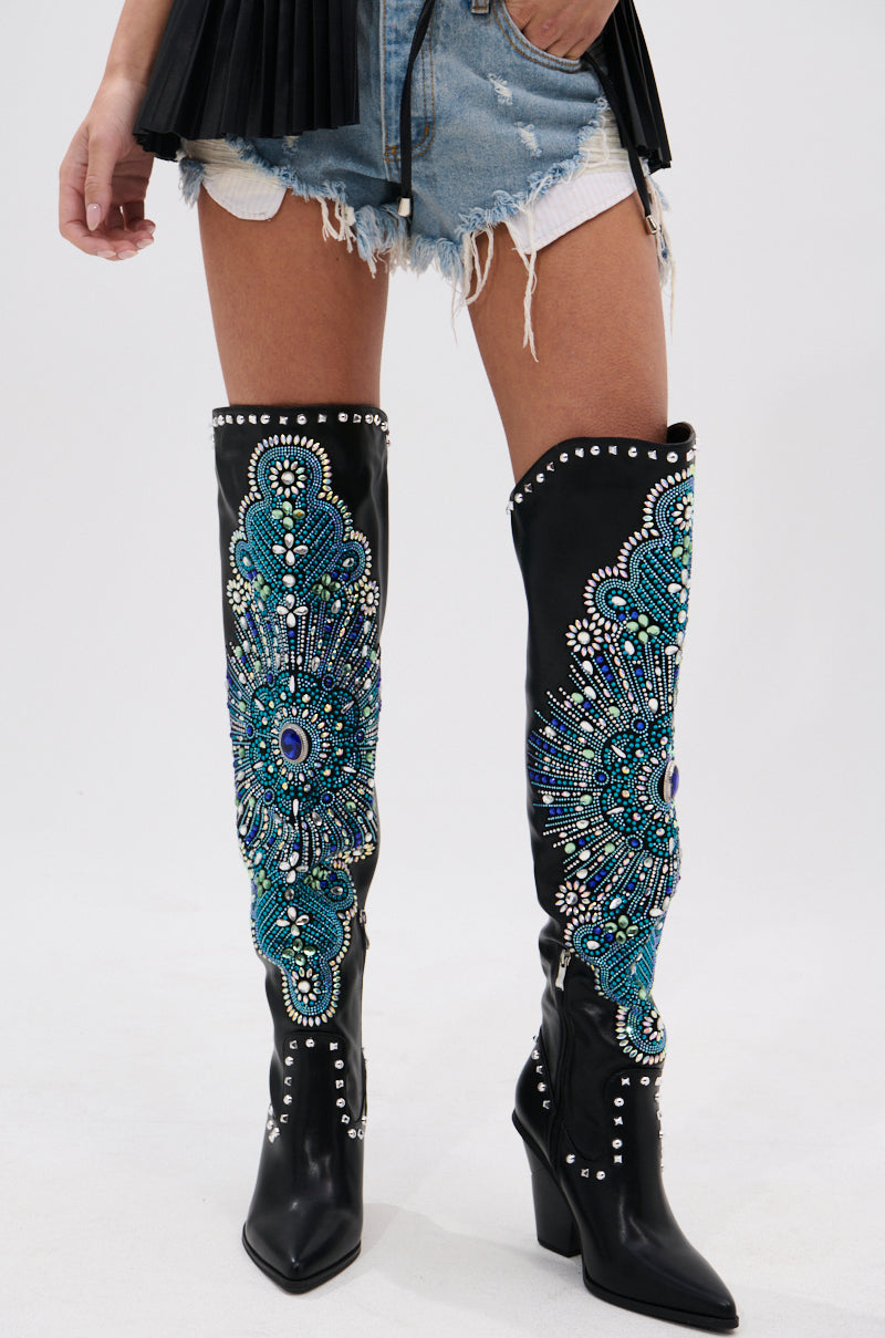 A person wears distressed denim shorts and AZALEA WANG AXELBEAT BLACK EMBELLISHED WESTERN BOOTS, featuring intricate blue, green, and silver rhinestone beadwork and stud accents, standing against a plain light background.