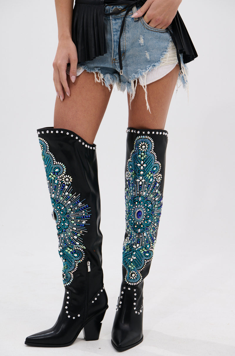 A person in distressed denim shorts, a black fringed belt, and AZALEA WANG AXELBEAT BLACK EMBELLISHED WESTERN BOOT with teal, blue, silver beads and metallic studs stands against a plain background.