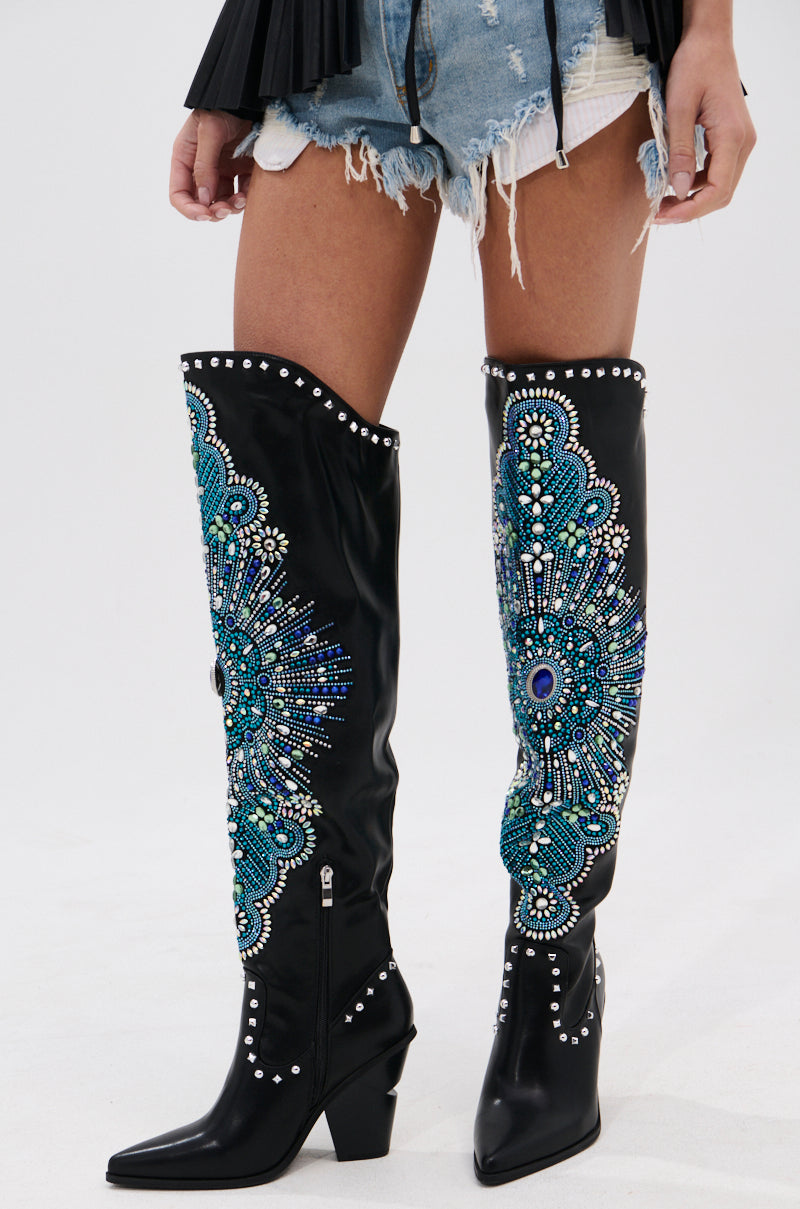 A person wearing distressed denim shorts and the AZALEA WANG AXELBEAT BLACK EMBELLISHED WESTERN BOOT, featuring intricate blue, green, and silver beadwork and studs, stands against a plain background.