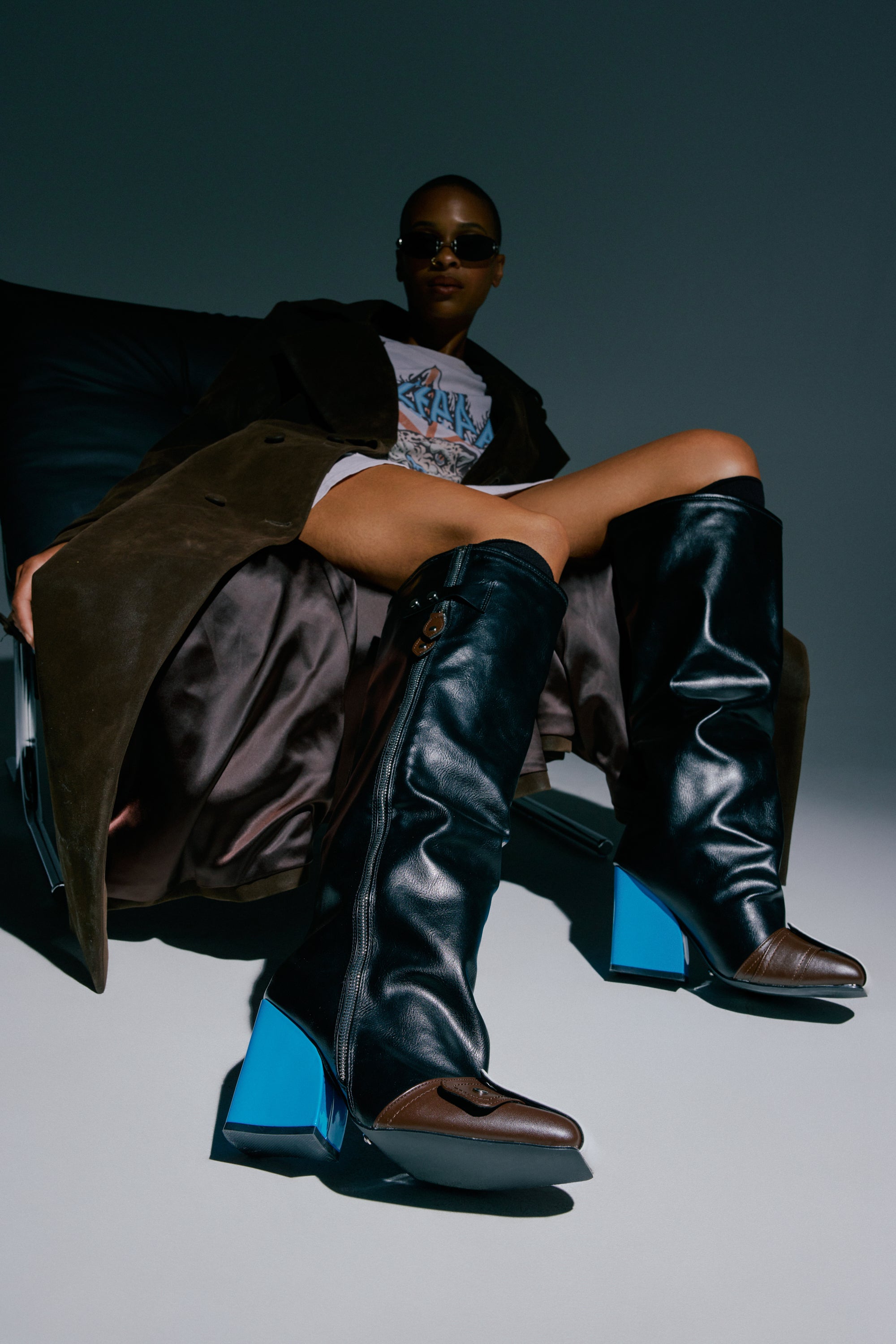 Wearing the AZALEA WANG AXTON BLACK MULTI COLOR BLOCKED BLOCK BOOT, a person with oversized boots featuring chunky blue heels sits in a chair, paired with sunglasses, a long dark coat, and a graphic shirt. Dramatic shadows and bold styling convey a striking modern fashion look.