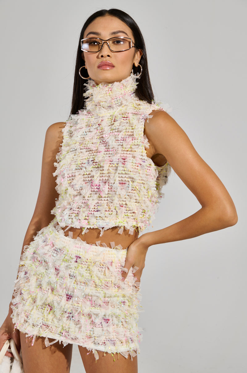 A person poses confidently in a fashionable ensemble featuring the SWEET LIKE CANDY SLEEVELESS TOP, crafted from textured, multicolored fabric. They wear glasses and have long hair, standing against a plain gray background.