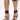 Wearing AZALEA WANG AYBARS Black Cuffed Sandals with silver western hardware, stiletto heels, studs, and wide ankle straps, the person carries a shiny black handbag against a plain white background.