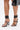Wearing AZALEA WANG AYBARS Black Cuffed Sandals with silver western hardware, stiletto heels, studs, and wide ankle straps, the person carries a shiny black handbag against a plain white background.