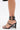 A close-up side view of the AZALEA WANG AYBARS BLACK CUFFED SANDAL WITH WESTERN HARDWARE, featuring stiletto heels and silver Western buckles—including an ankle buckle—worn by a person against a plain white background.