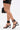 A person walks on a plain white background wearing a short black skirt and AZALEA WANG AYBARS Black Cuffed Sandals with Western Hardware, carrying a black handbag.