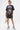 A woman with a short hairstyle and leg tattoos stands against a white background wearing the AALIYAH FOREVER GRAPHIC TSHIRT DRESS, paired with a sparkly silver handbag and fluffy blue slippers.