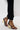A person stands on a white surface wearing black pants and the AZALEA WANG AYYAR BLACK GOLD WEDGE HEEL SANDAL with gold accents; part of a white garment's hem is also visible.
