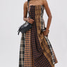 A woman poses against a white backdrop in the FALL IN LOVE PLAID MAXI DRESS, featuring 90’s-inspired brown and black plaid, styled with a black crossbody bag and studded shoes for edgy punk princess vibes.