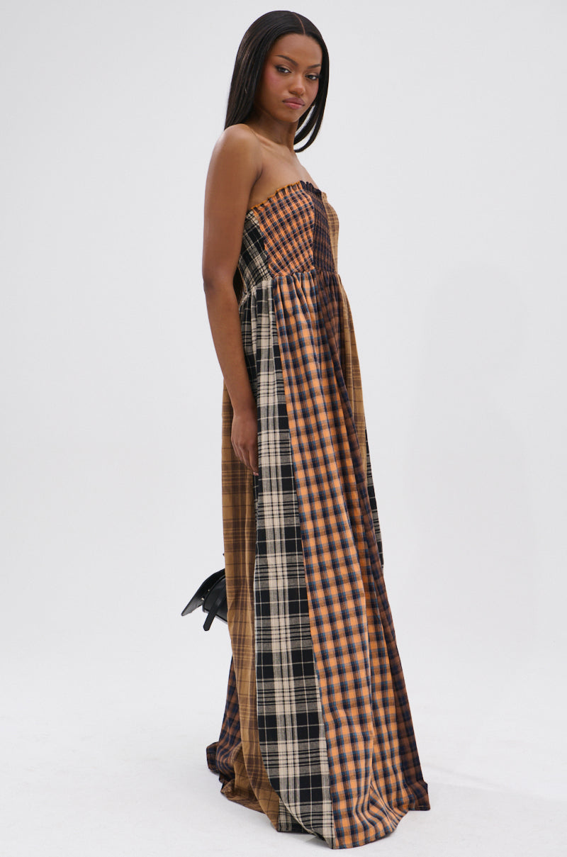 A woman models the FALL IN LOVE PLAID MAXI DRESS, a 90’s-inspired strapless, floor-length gown in brown, black, and beige plaid. She stands against a white background, channeling subtle punk princess vibes as she looks to the side.