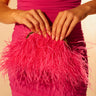A woman in a fitted, ruched hot pink dress holds the TICKLED PINK PURSE, its vibrant fuchsia feathers and lustrous hot pink satin softly glowing as she poses against a gently lit background.