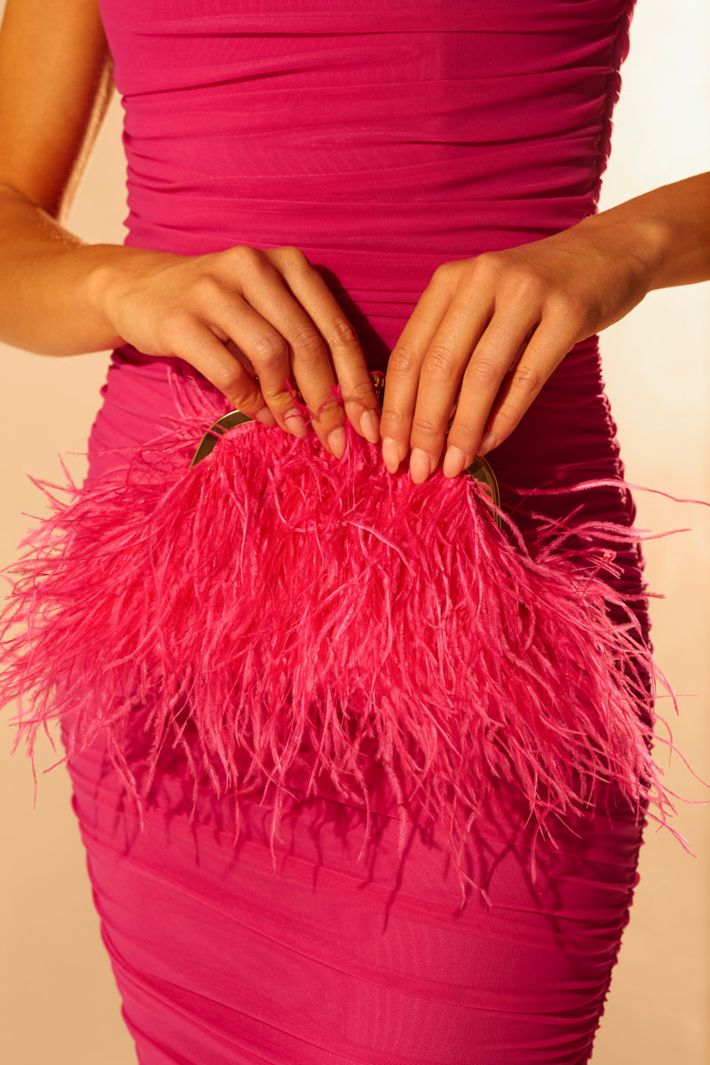A woman in a fitted, ruched hot pink dress holds the TICKLED PINK PURSE, its vibrant fuchsia feathers and lustrous hot pink satin softly glowing as she poses against a gently lit background. 0