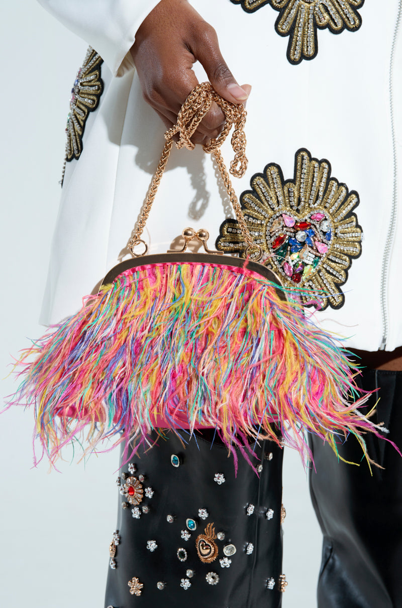 A person holds the AZONTINA PINK FEATHER PURSE, a mini bag embellished with colorful feathers, including a striking pink feather, and a gold chain strap. They wear a white jacket with embroidered floral patterns and black pants decorated with multicolored embellishments.