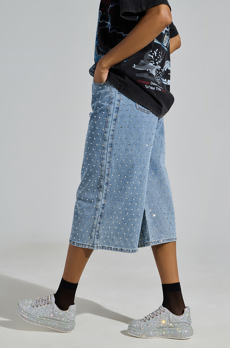 A person wears a black shirt with long, light blue denim shorts featuring white polka dots, and AZALEA WANG AZURE SILVER RHINESTONE SNEAKERS with black socks against a simple gray backdrop.
