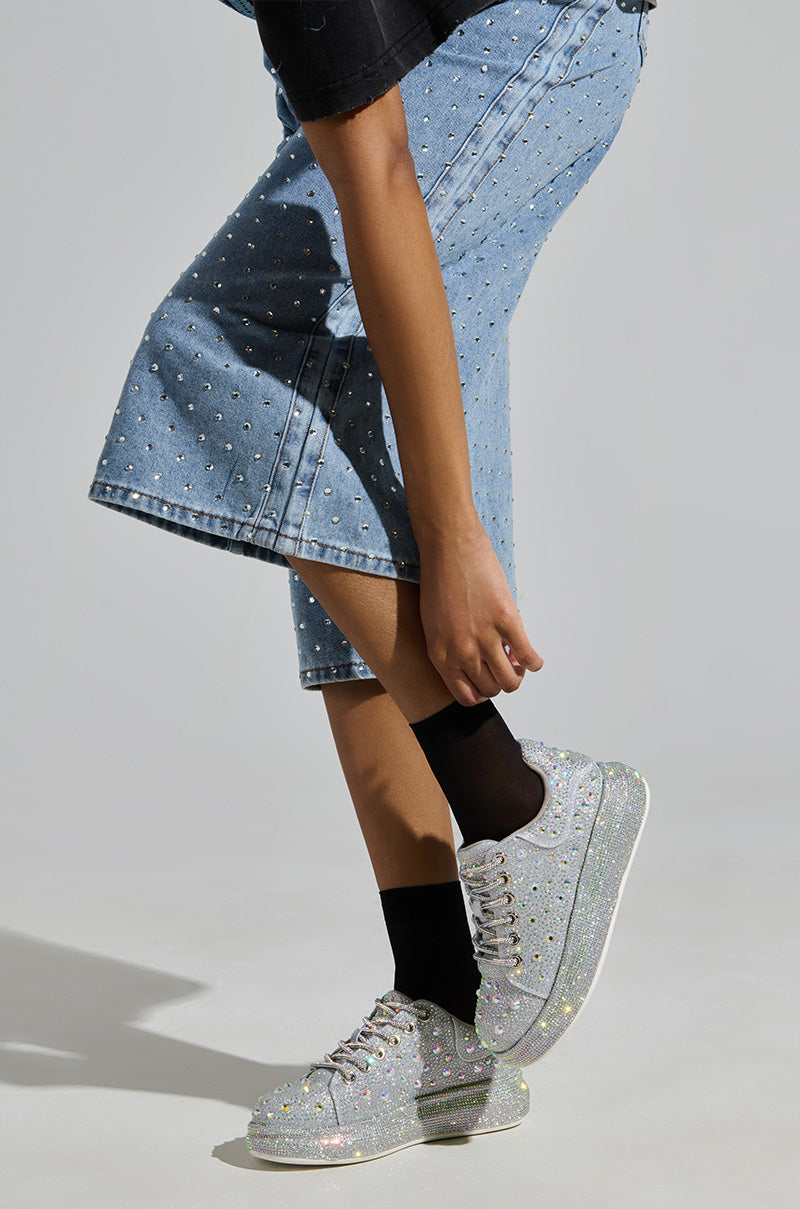 Wearing denim pants with rhinestone embellishments, sparkling AZALEA WANG AZURE SILVER RHINESTONE SNEAKERS, and black socks, a person is slightly bent forward, their left hand touching the shorts' edge against a plain light gray background.