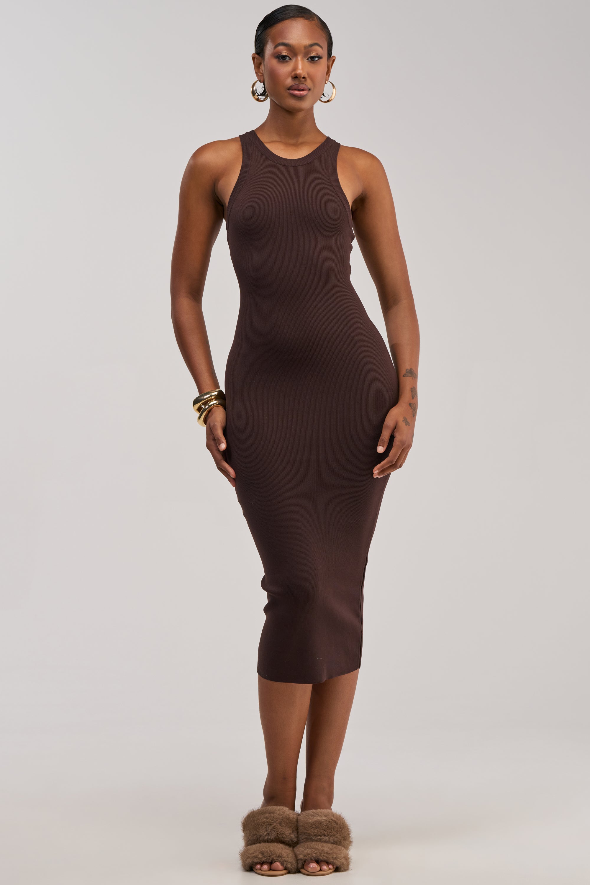 A woman wearing the CUTIE PIE KNIT MIDI DRESS IN BROWN, paired with gold hoop earrings, bracelets, and brown fuzzy sandals, stands against a plain light background.