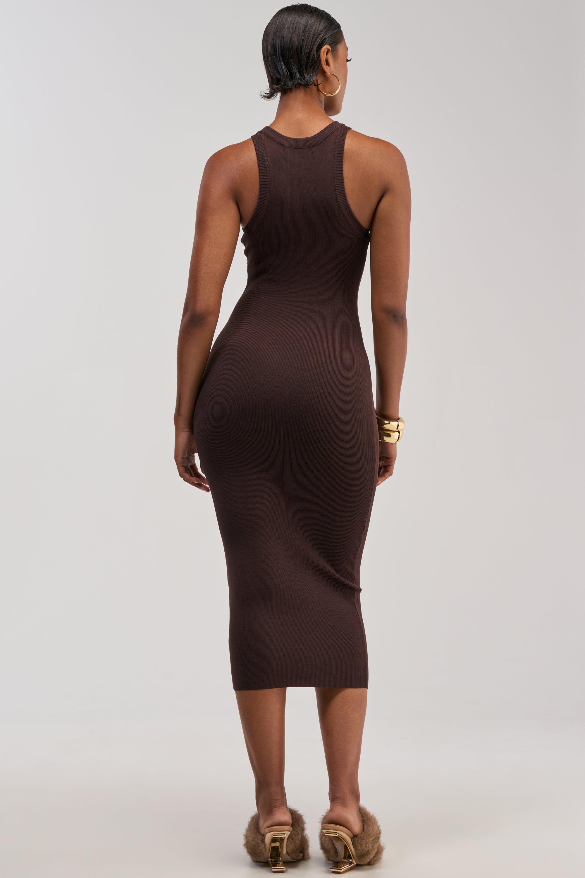 A woman faces away, wearing the CUTIE PIE KNIT MIDI DRESS IN BROWN—a fitted, sleeveless knit tank midi dress—paired with fur-trimmed heels. She styles her short hair with gold hoop earrings and chunky gold bracelets, set against a plain, light-colored background.
