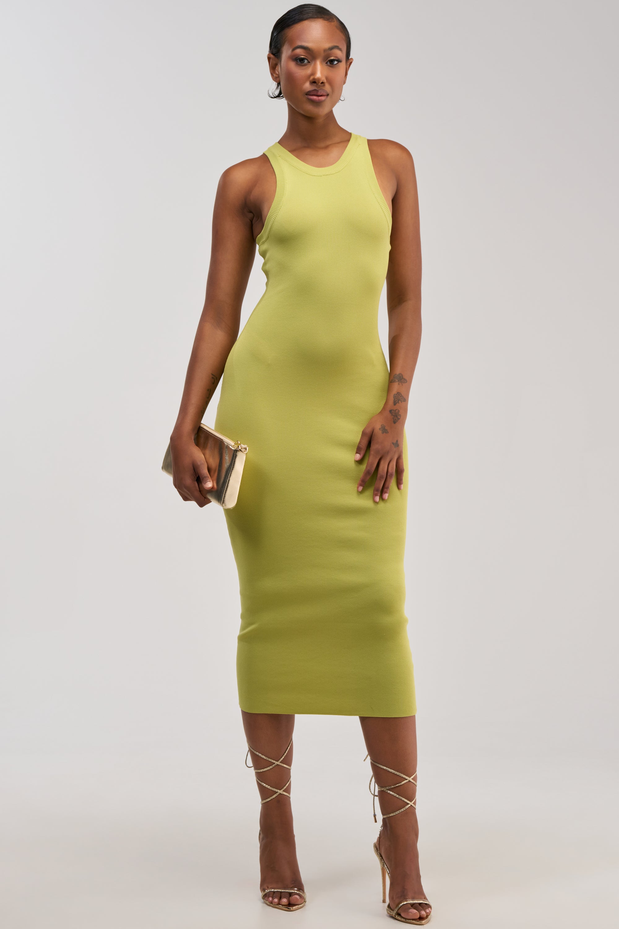 A woman wearing the CUTIE PIE KNIT MIDI DRESS IN CHARTREUSE and strappy high heels, holding a gold clutch, stands against a plain white background.