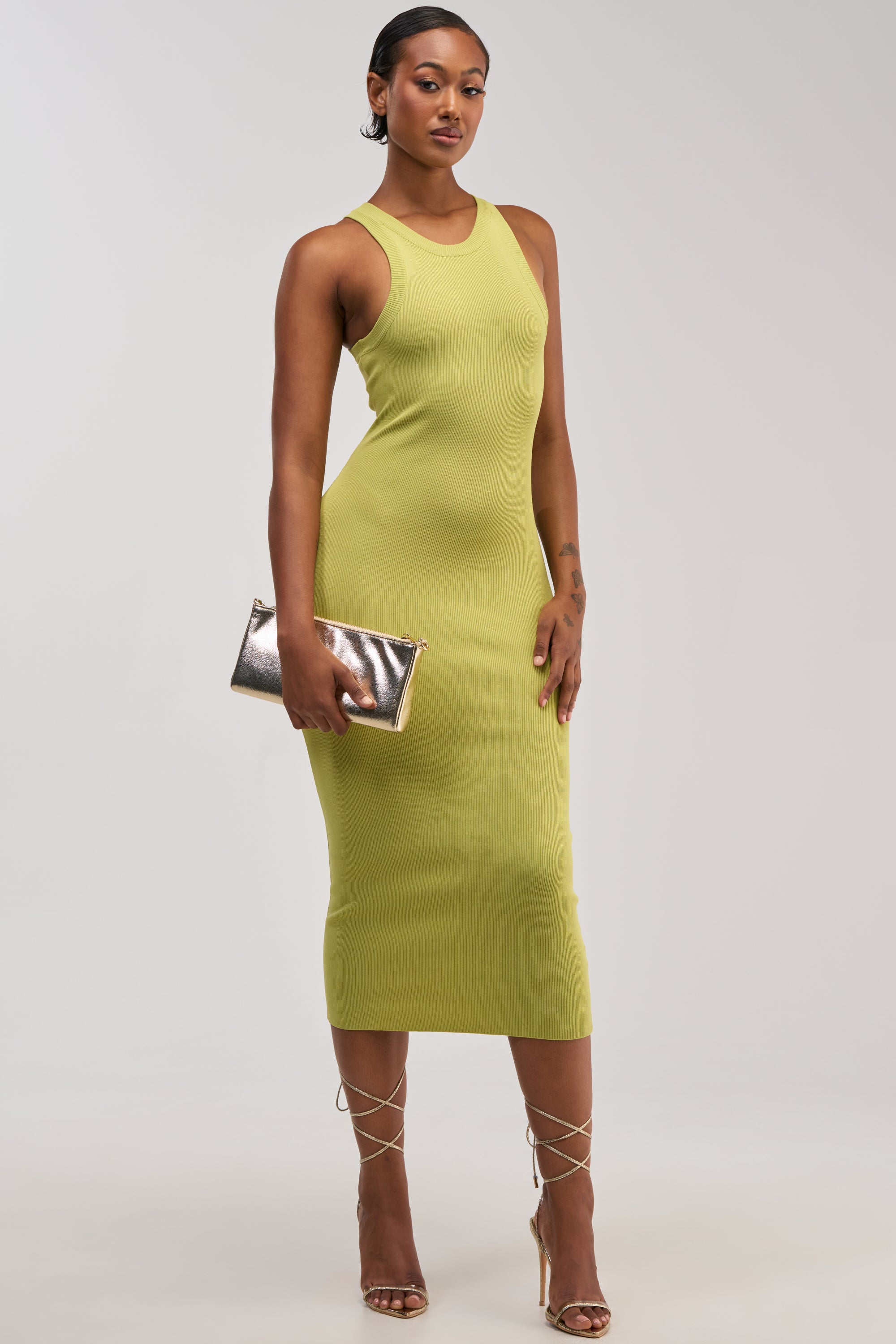 Dressed in the “CUTIE PIE KNIT MIDI DRESS IN CHARTRUESE,” a woman poses against a plain light background, holding a metallic clutch and wearing strappy heels. The sleeveless, figure-hugging knit dress features a fresh chartreuse hue for an effortlessly chic look.