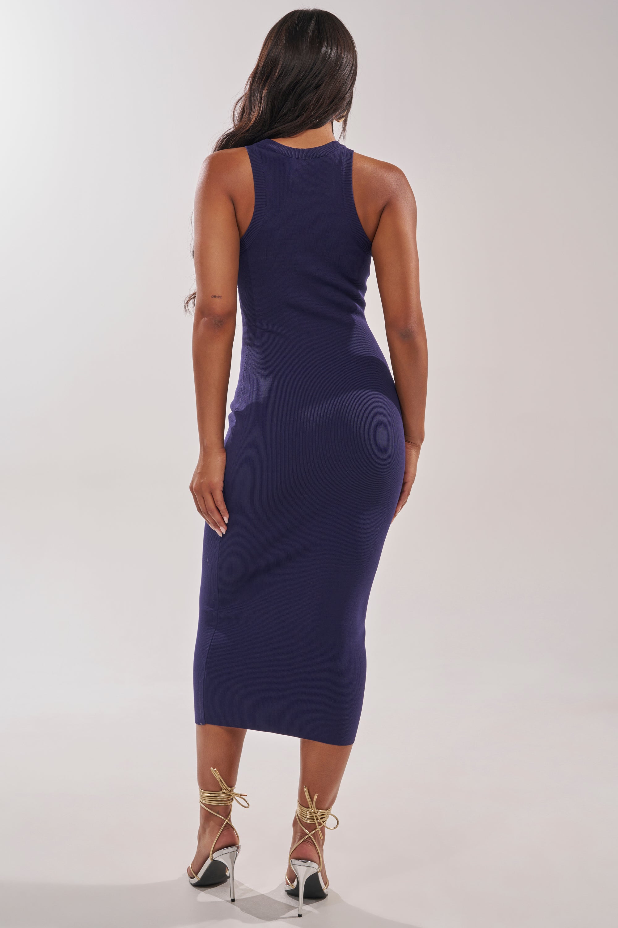 A woman with long, wavy hair stands with her back to the camera, wearing the CUTIE PIE KNIT MIDI DRESS IN NAVY and white ankle-strap high-heeled sandals against a plain light background. 3